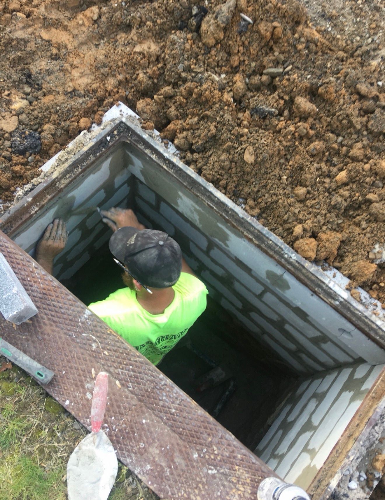 A worker in a bright yellow shirt uses a trowel to apply mortar to the concrete block wall of an open underground trench.
