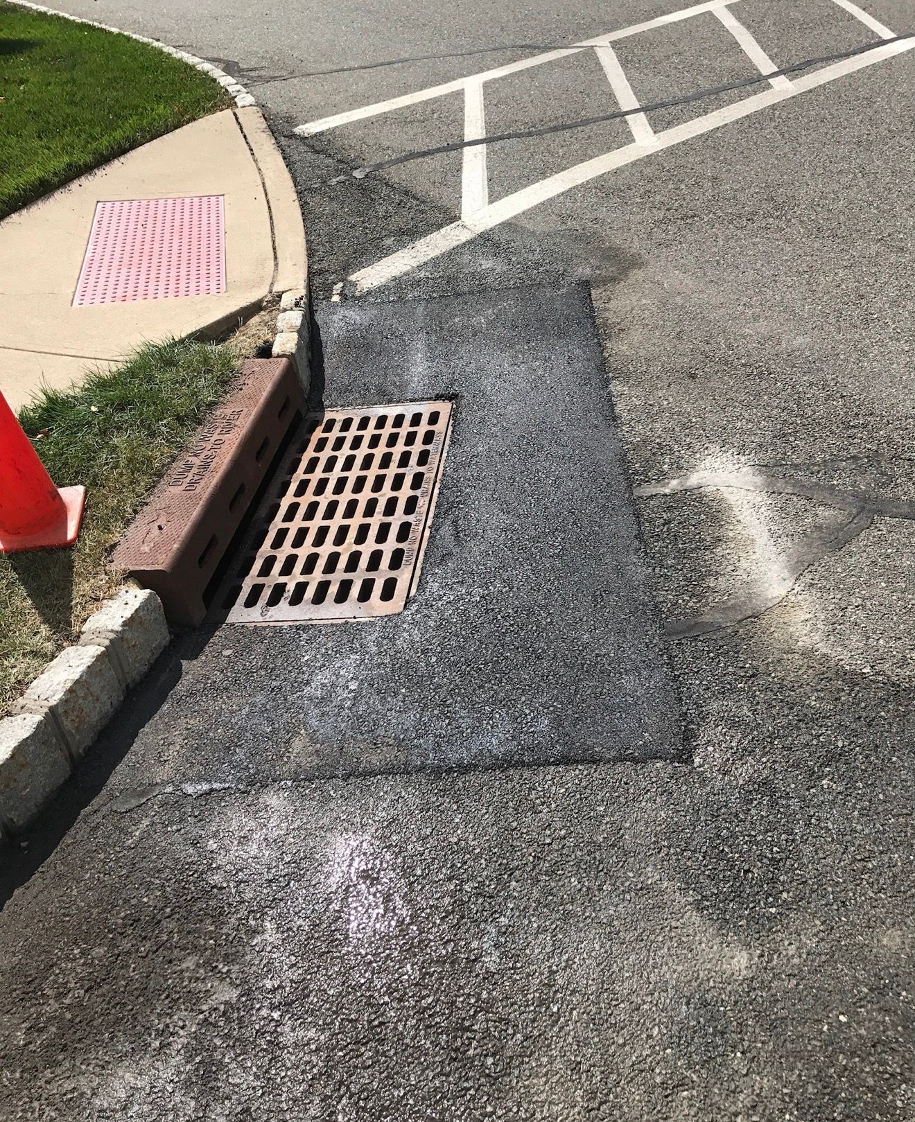 A metal storm drain grate set into fresh asphalt at a street corner next to a sidewalk with a red traffic cone nearby.