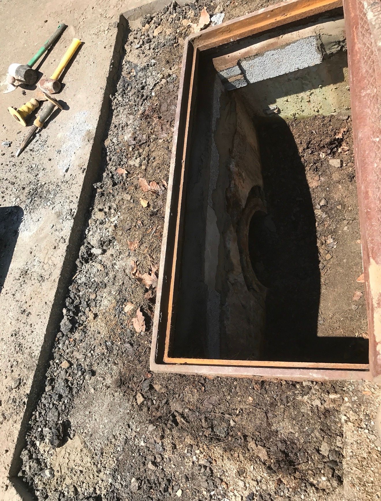 An open rectangular utility access hole in the ground, with tools laid out on the dirt nearby.
