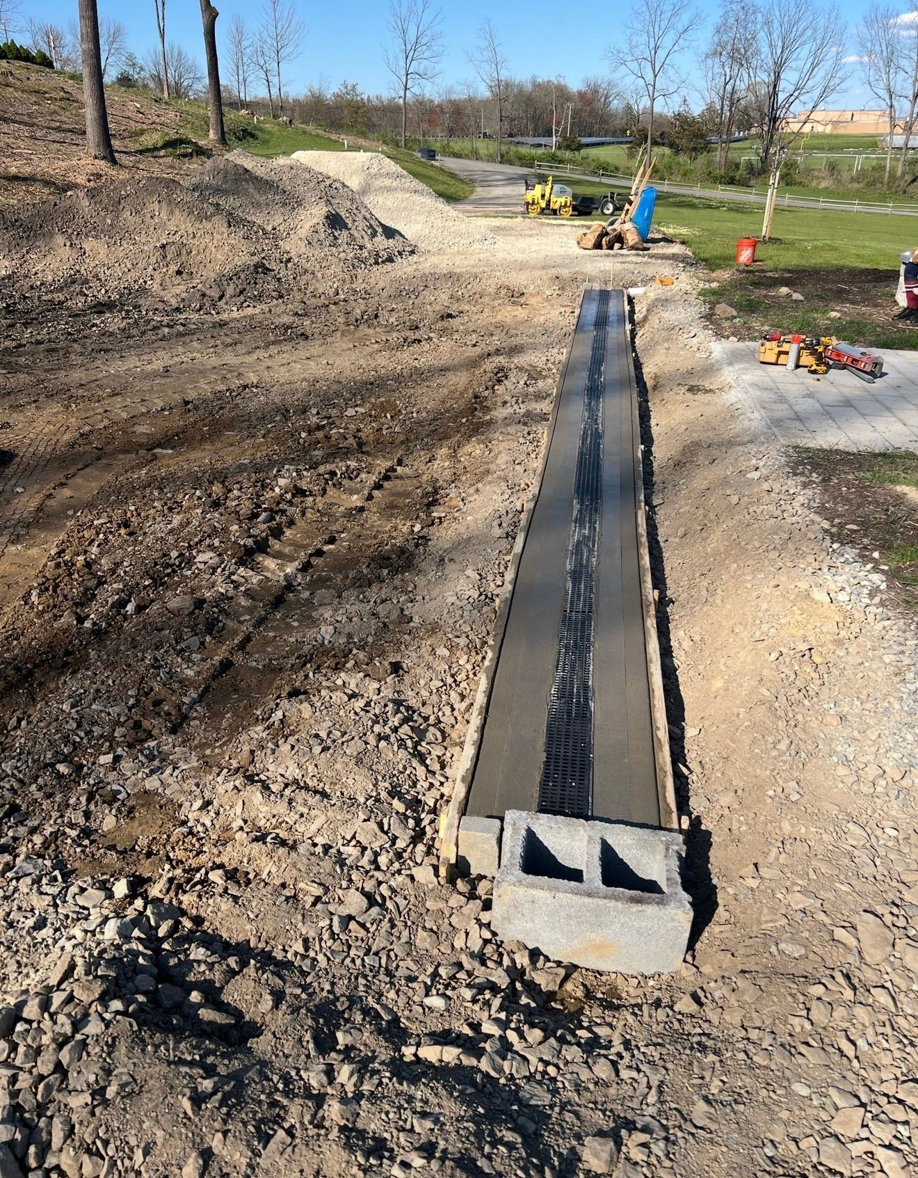 A concrete footing and a single cinder block sit on a dirt construction site, with a dark strip running along the center.