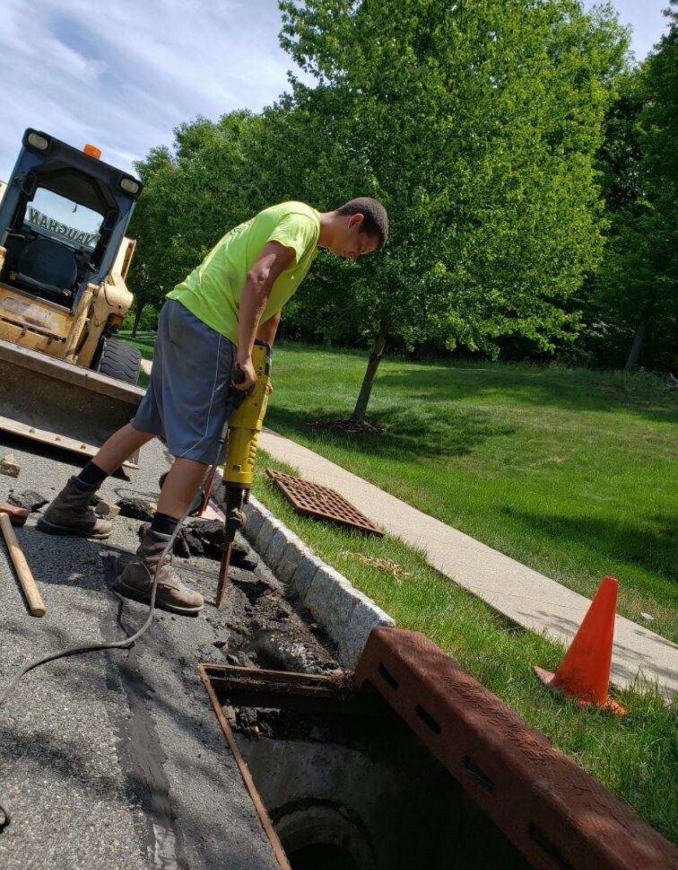 A worker in a high-visibility yellow shirt uses a jackhammer to break up asphalt next to a drainage grate and traffic cone.