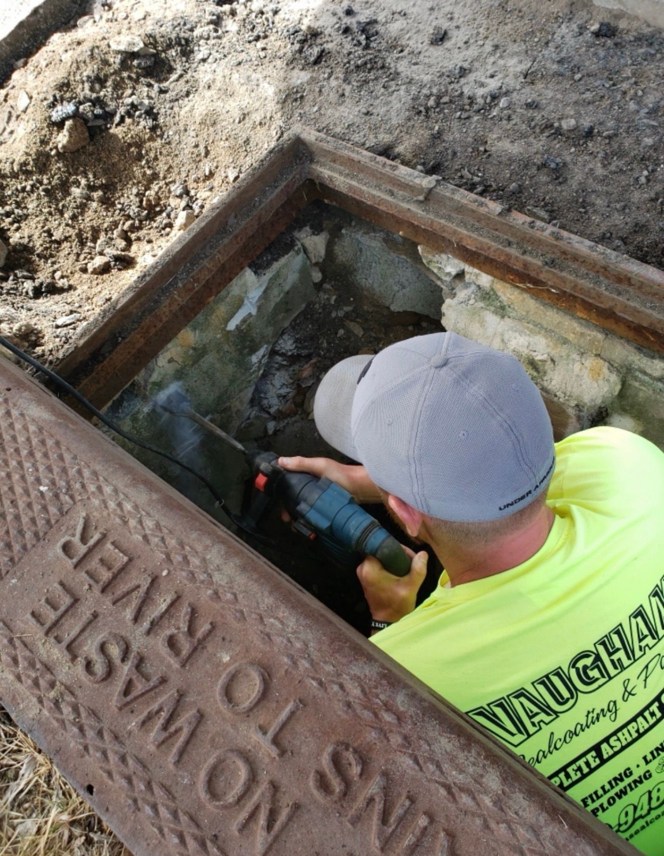 A worker in a neon yellow shirt uses a tool to perform maintenance inside an open roadside storm drain.