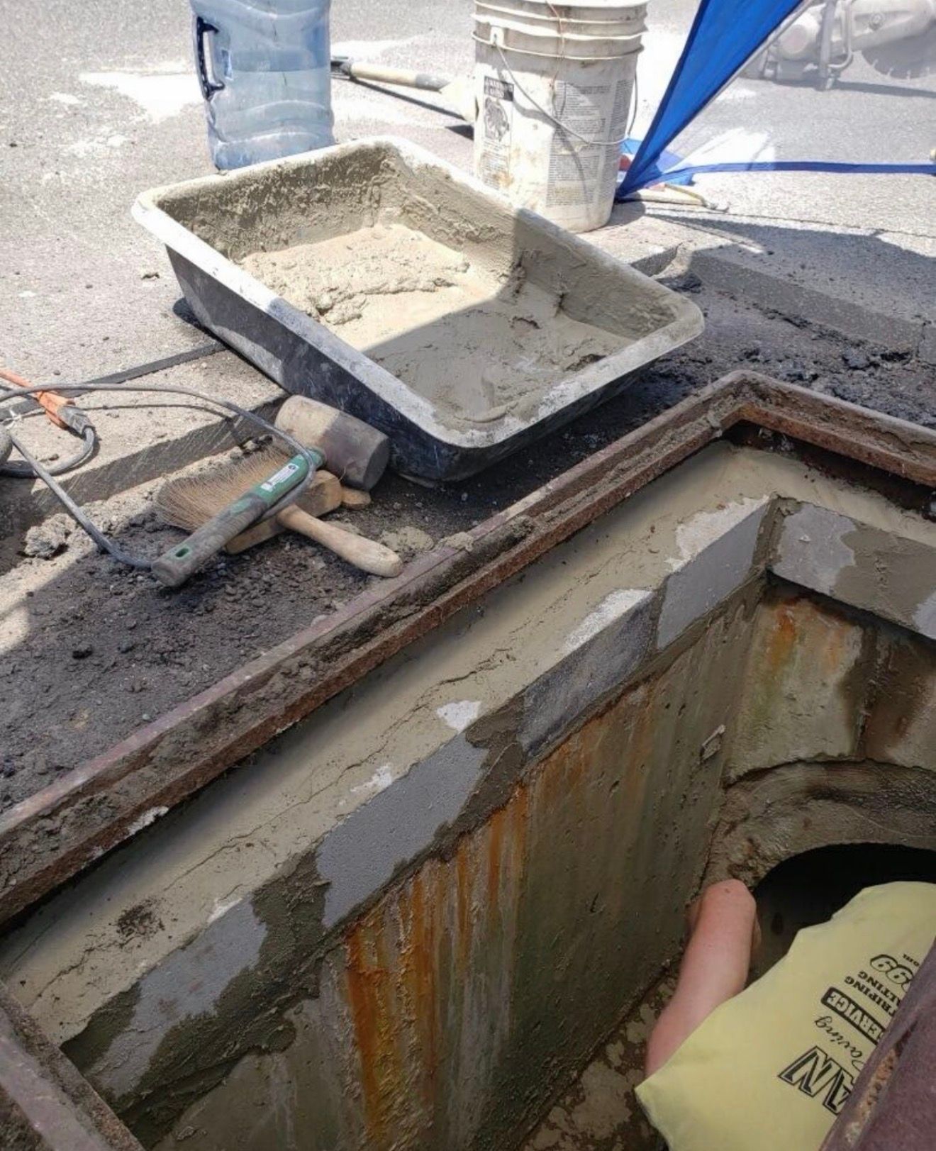 A worker in a yellow shirt repairs a concrete utility hole, with a mortar tray and bucket nearby on the pavement.