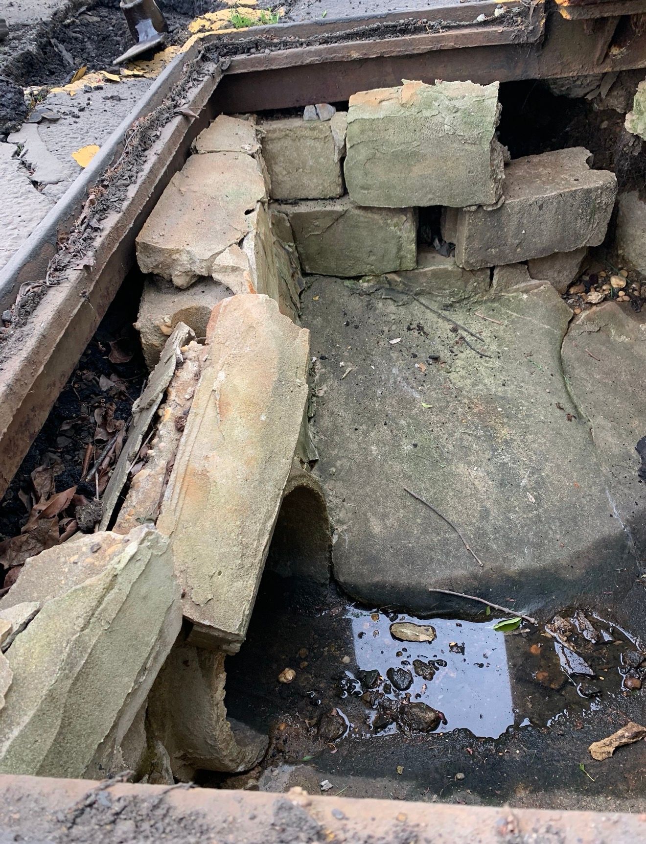 A top-down view of a crumbling concrete storm drain catch basin with stagnant water and scattered debris.
