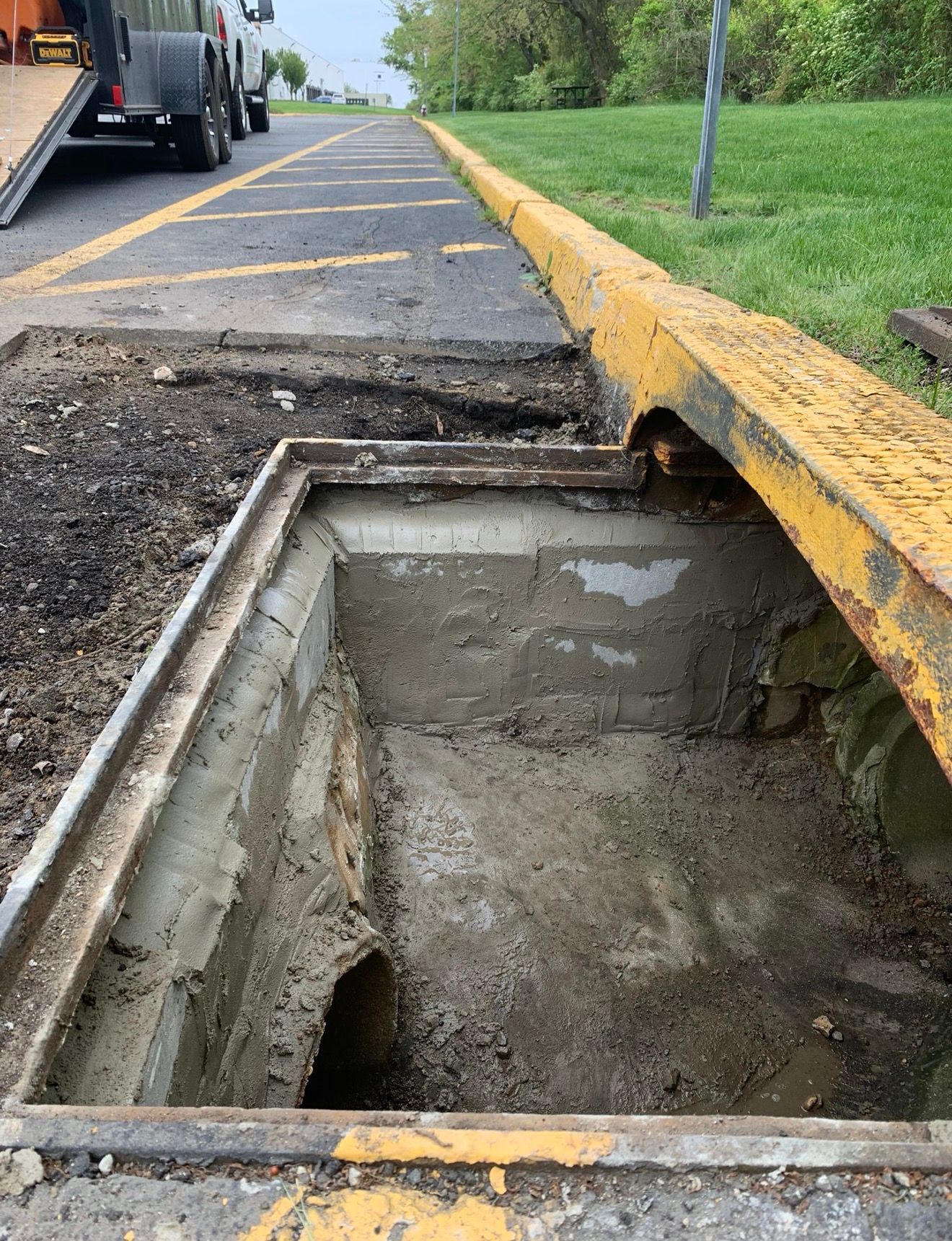 An open concrete storm drain box sits along a yellow curb next to a parking lot area.