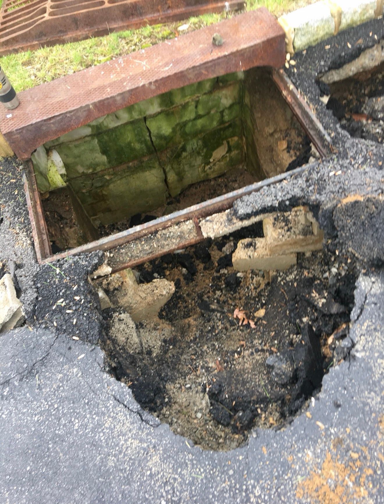 A damaged concrete storm drain with a missing cover and broken asphalt surface around the opening.