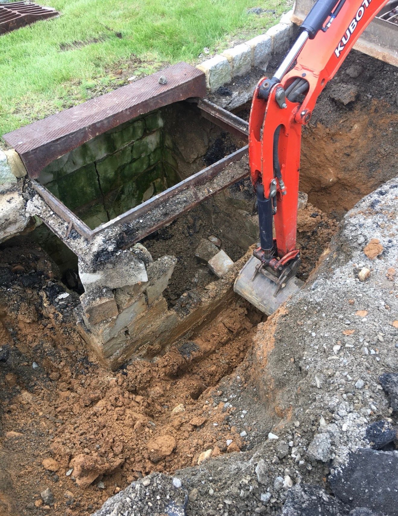 An orange excavator arm digs next to a brick-lined utility vault in a construction trench.