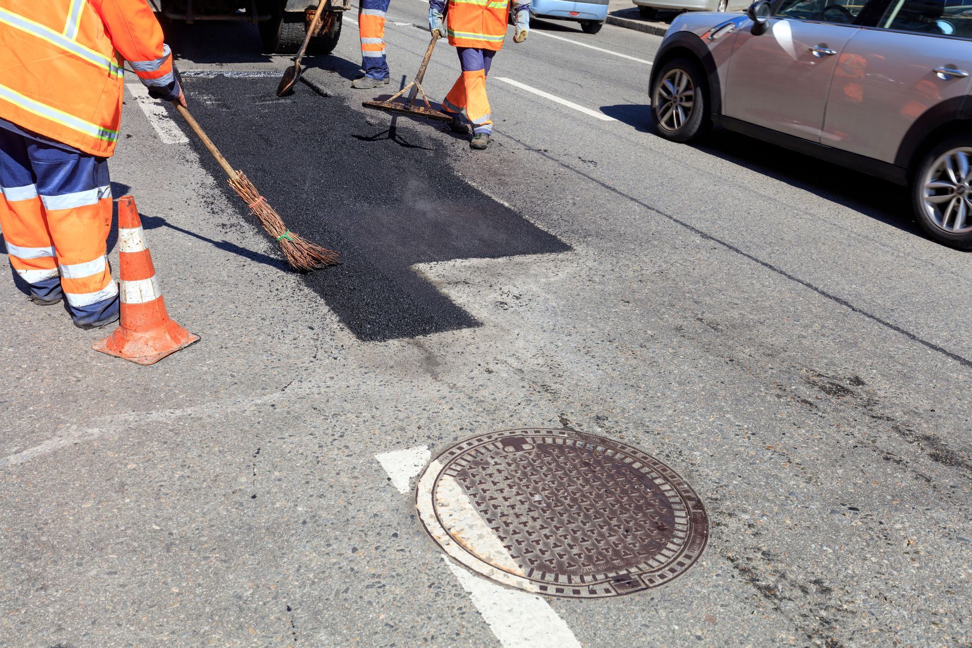 Workers in high-visibility orange gear use rakes to spread fresh asphalt on a sunny road near a manhole cover.