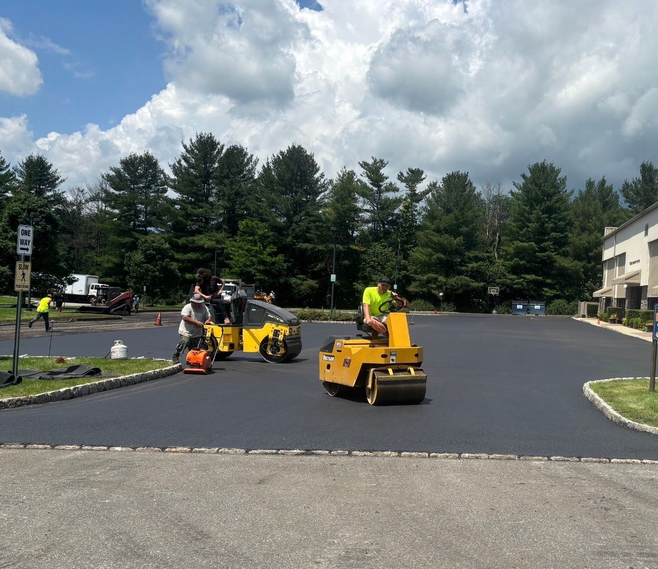 Two workers operate yellow ride-on rollers to smooth fresh black asphalt in a parking lot under a cloudy sky.