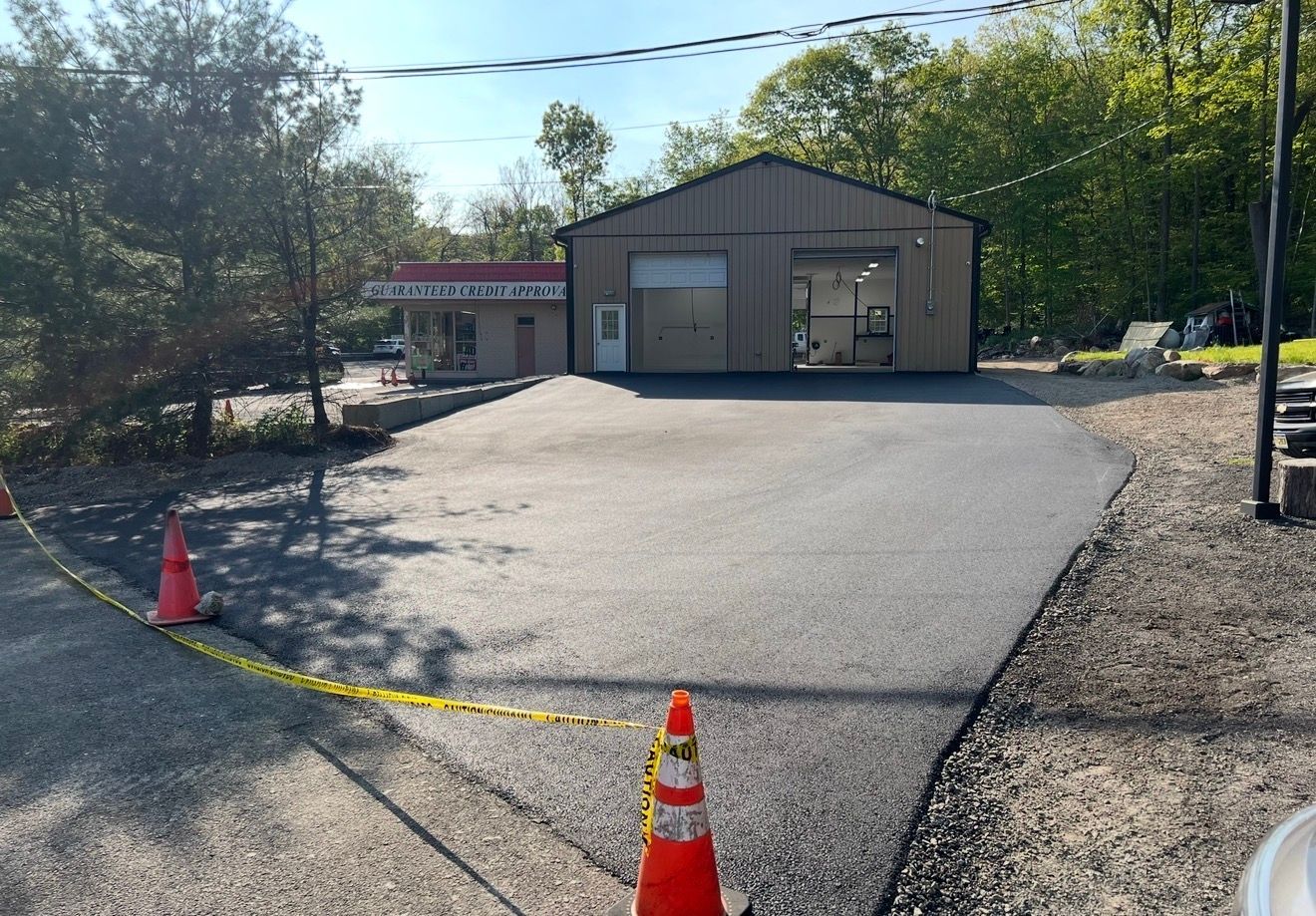 A newly paved asphalt driveway leads to a metal workshop building on a sunny day, marked by orange construction cones.