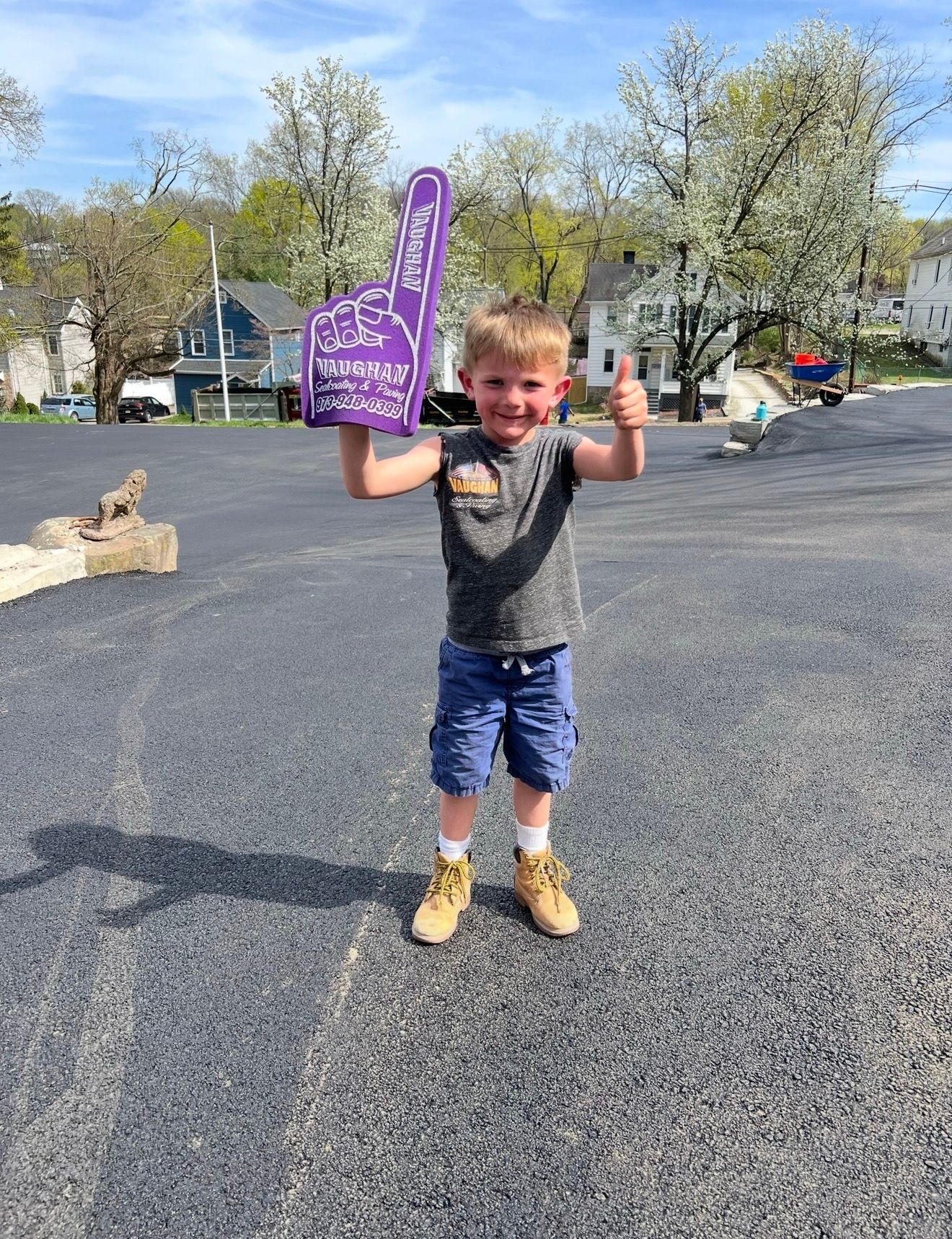 A child in a grey shirt and blue shorts stands on an asphalt lot, smiling, giving a thumbs-up and holding a purple foam hand.