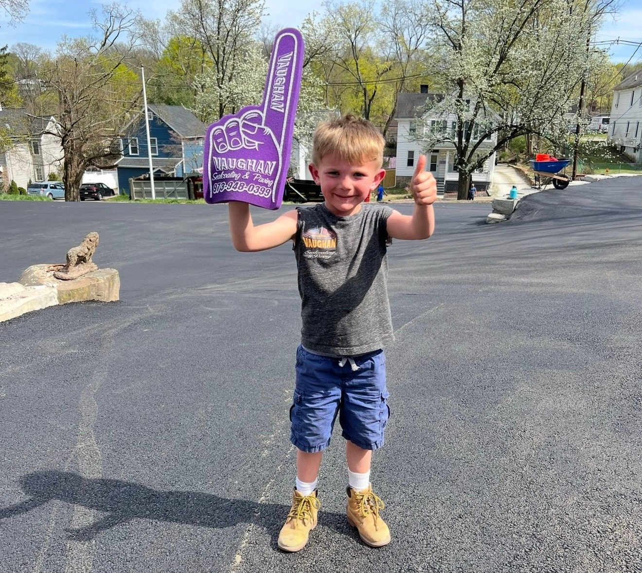 A child stands on an asphalt lot, holding a purple foam finger while giving a thumbs-up.