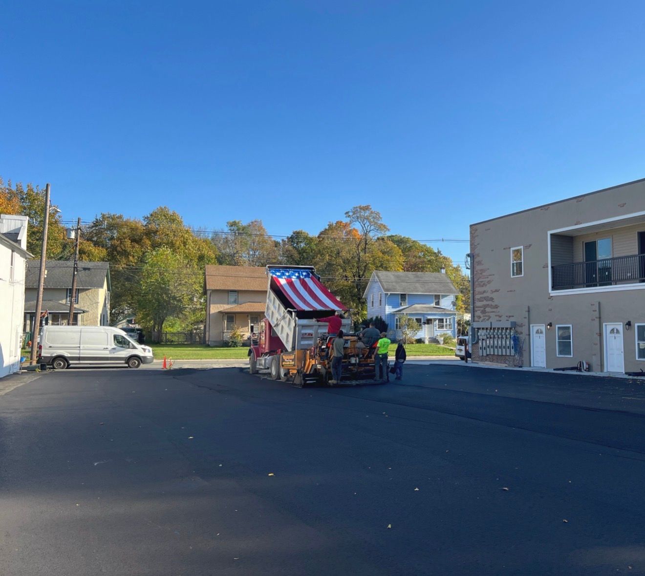 Workers pave a parking lot with a red, white, and blue truck parked in the center on a sunny day.