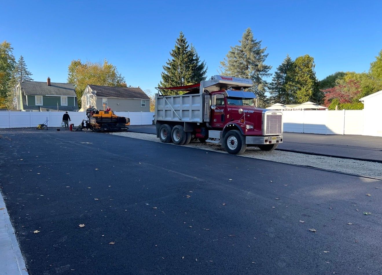 A red dump truck sits on a newly paved asphalt surface next to a construction vehicle in a sunny suburban neighborhood.