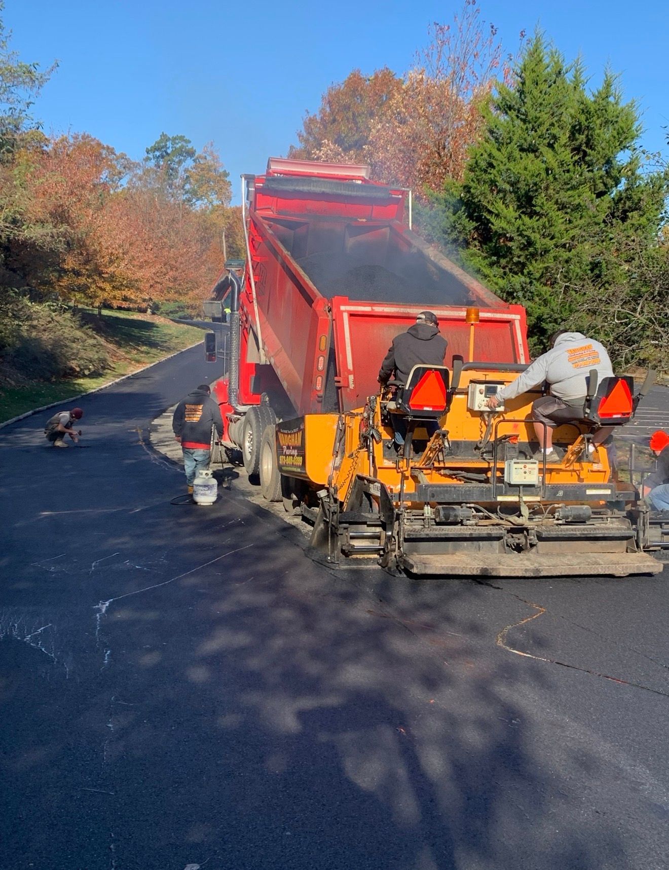 A dump truck fills an orange asphalt paver on a road with autumn trees in the background as workers oversee the operation.