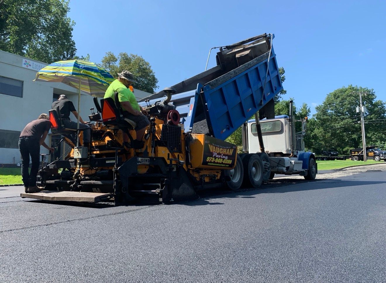 A dump truck deposits asphalt into a yellow paving machine on a sunny residential street as workers operate the equipment.