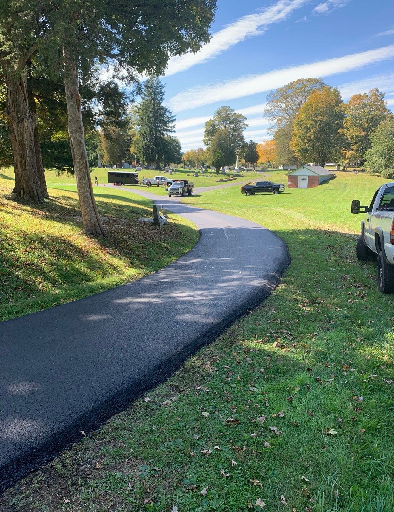 A newly paved asphalt path curves through a grassy park area on a sunny day with vehicles parked in the background.