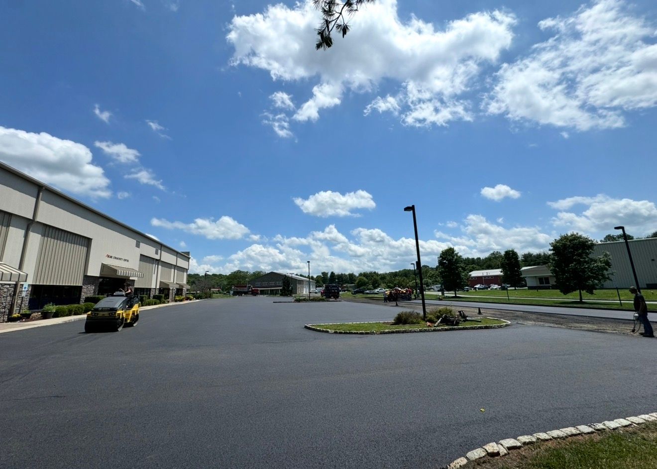 A bright outdoor scene of a large, empty asphalt parking lot next to a warehouse building under a blue, cloudy sky.