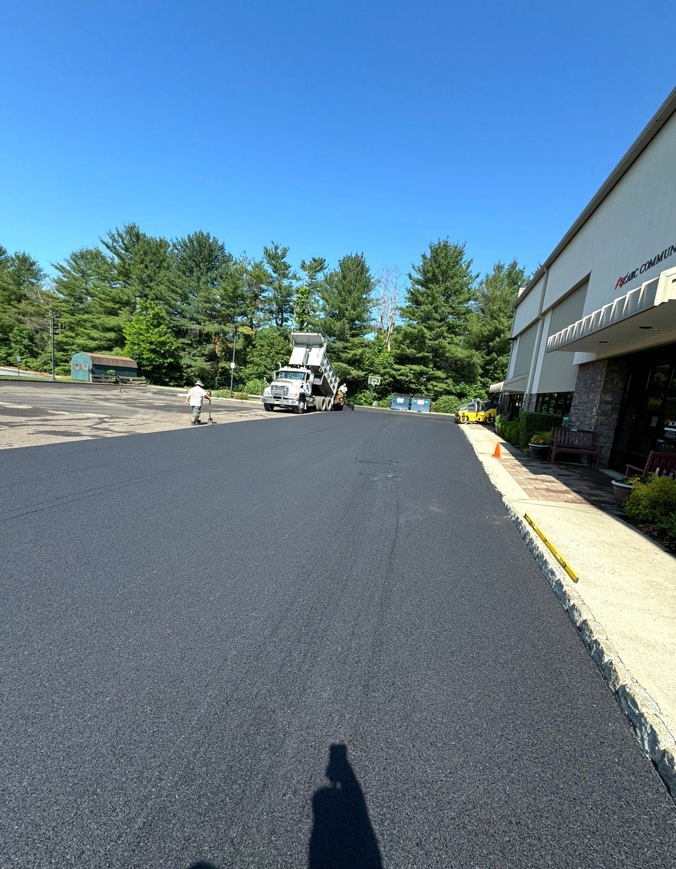 Freshly paved asphalt surface leading toward a construction vehicle in a sunny parking lot next to a building.