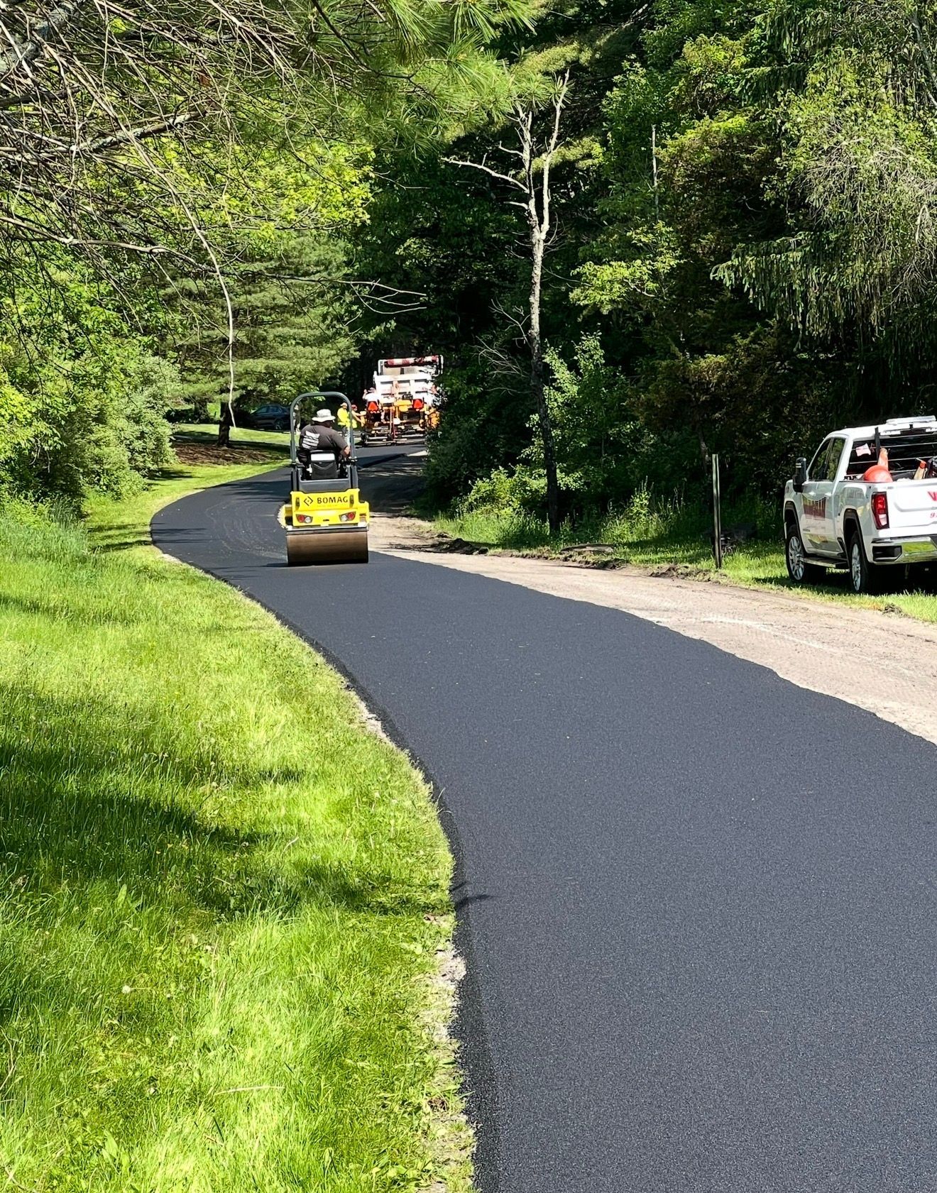 A yellow road roller paves a fresh black asphalt path through a green, wooded area.