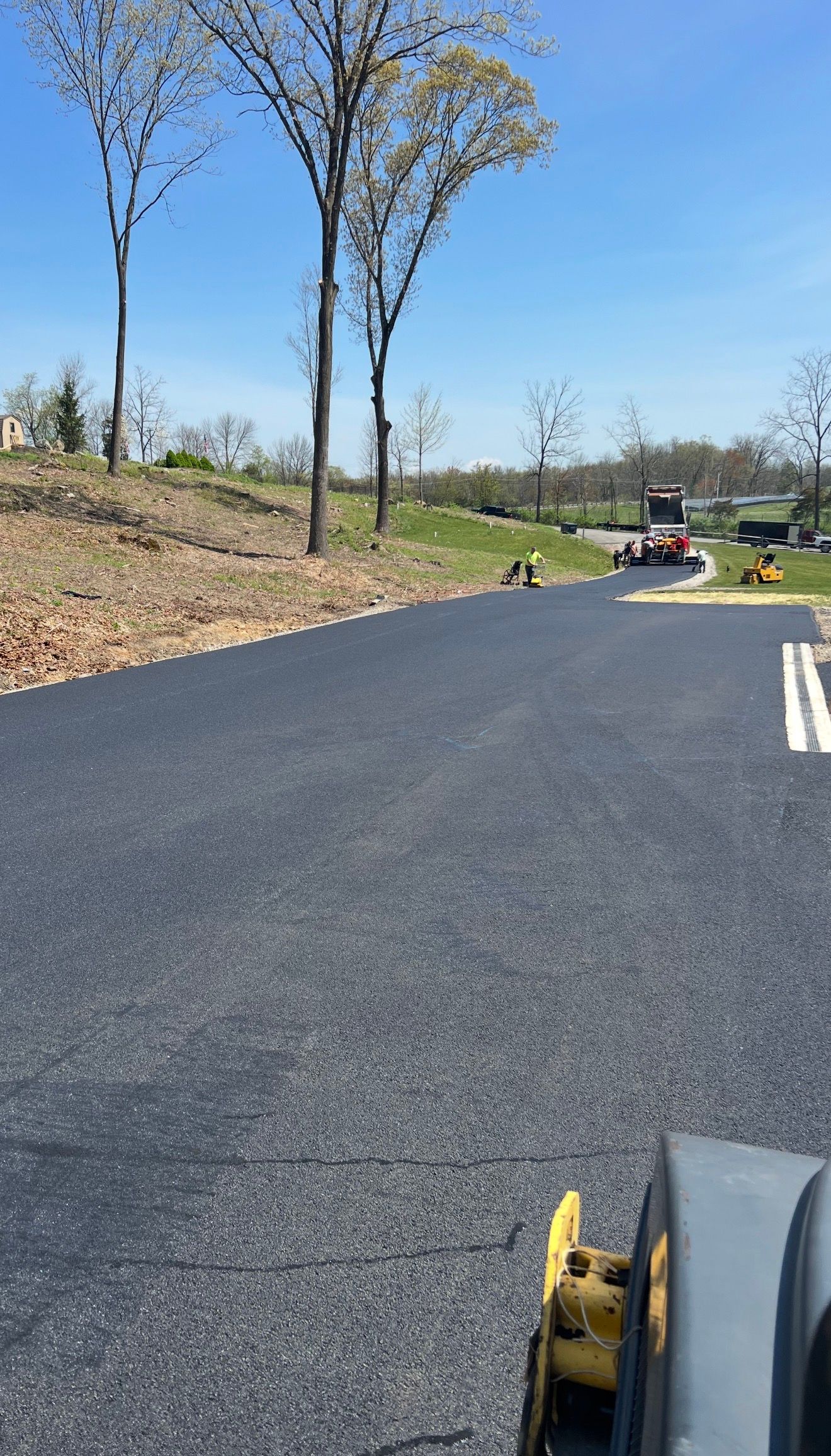 Fresh asphalt paving on a new road under a blue sky, with heavy machinery visible in the distance.