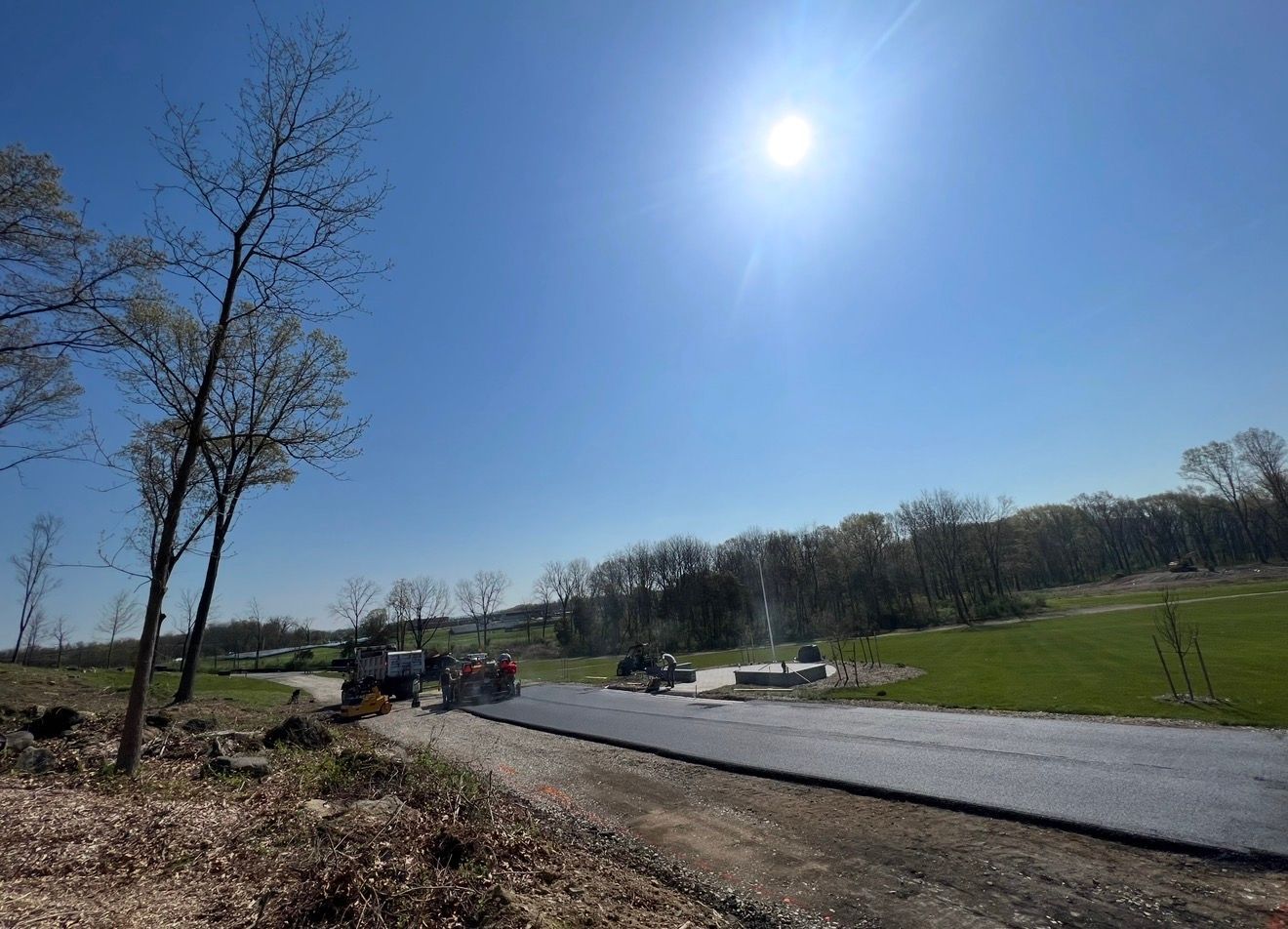 A construction crew paving a road through a rural, sunny landscape with trees and grass.