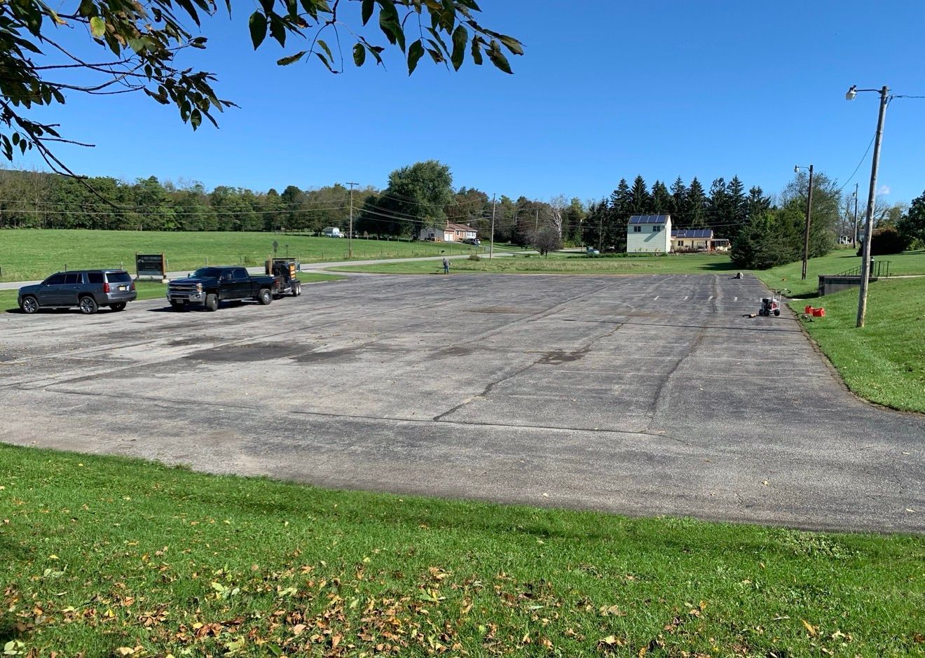 An asphalt parking lot sits on a grassy field under a clear blue sky, with two parked pickup trucks on the left.