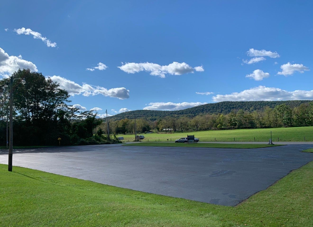 A newly paved black asphalt lot bordered by green grass, with trees and a mountain ridge under a bright blue sky.