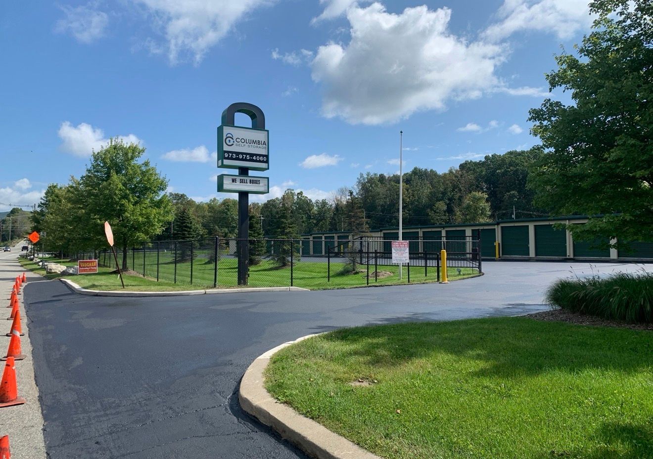 A storage facility with a tall sign, paved driveway, green lawns, and orange safety cones under a partly cloudy sky.