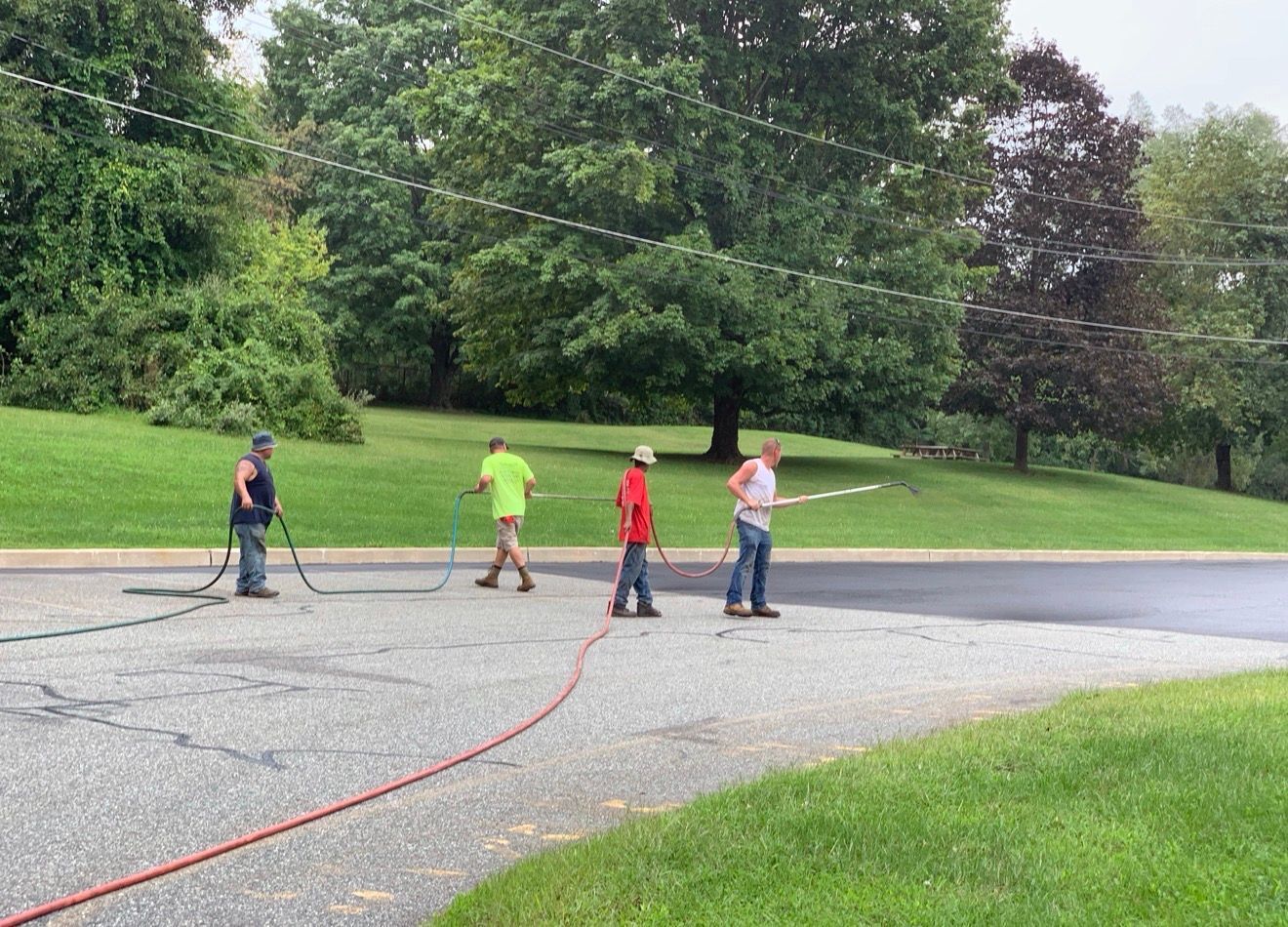 Four workers in safety vests apply crack sealer to a residential asphalt driveway, stretching a hose across the surface.