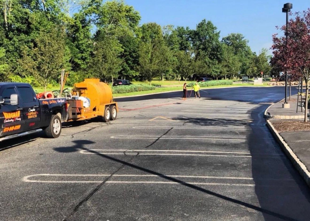 A truck towing an orange pavement sealcoating tank in a parking lot, with two workers in the background.
