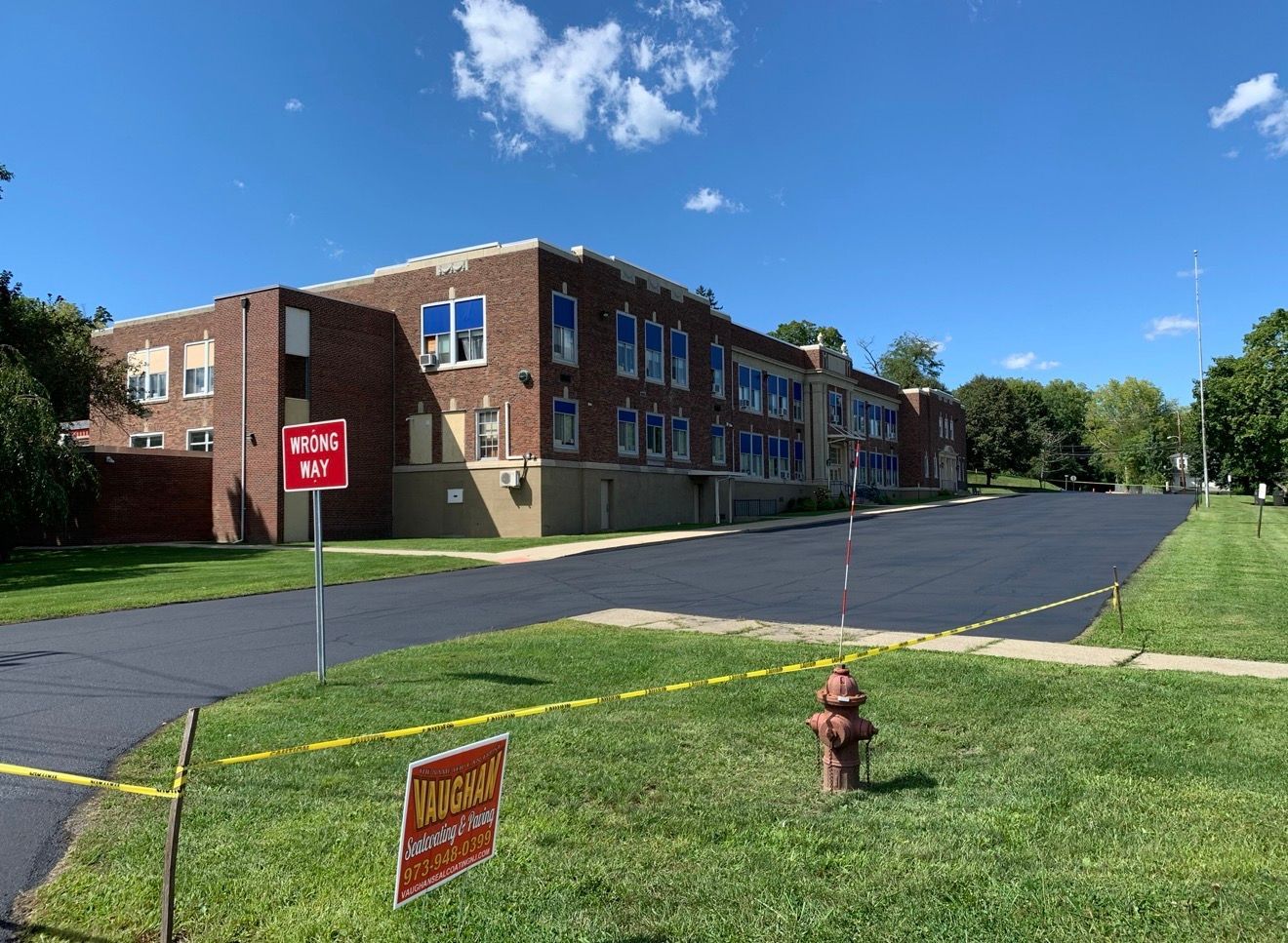 A large brick school building with a newly paved parking lot in front, a fire hydrant, and caution tape on a sunny day.