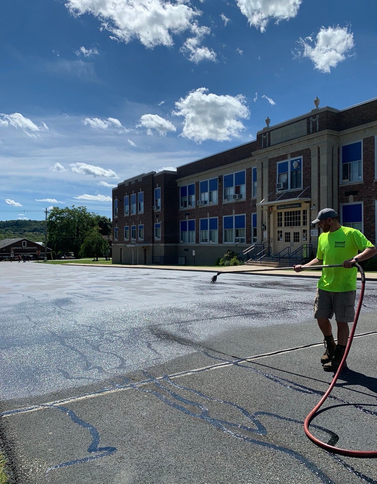 A worker in a neon green shirt uses a sprayer to coat a dark asphalt parking lot in front of a brick school building.