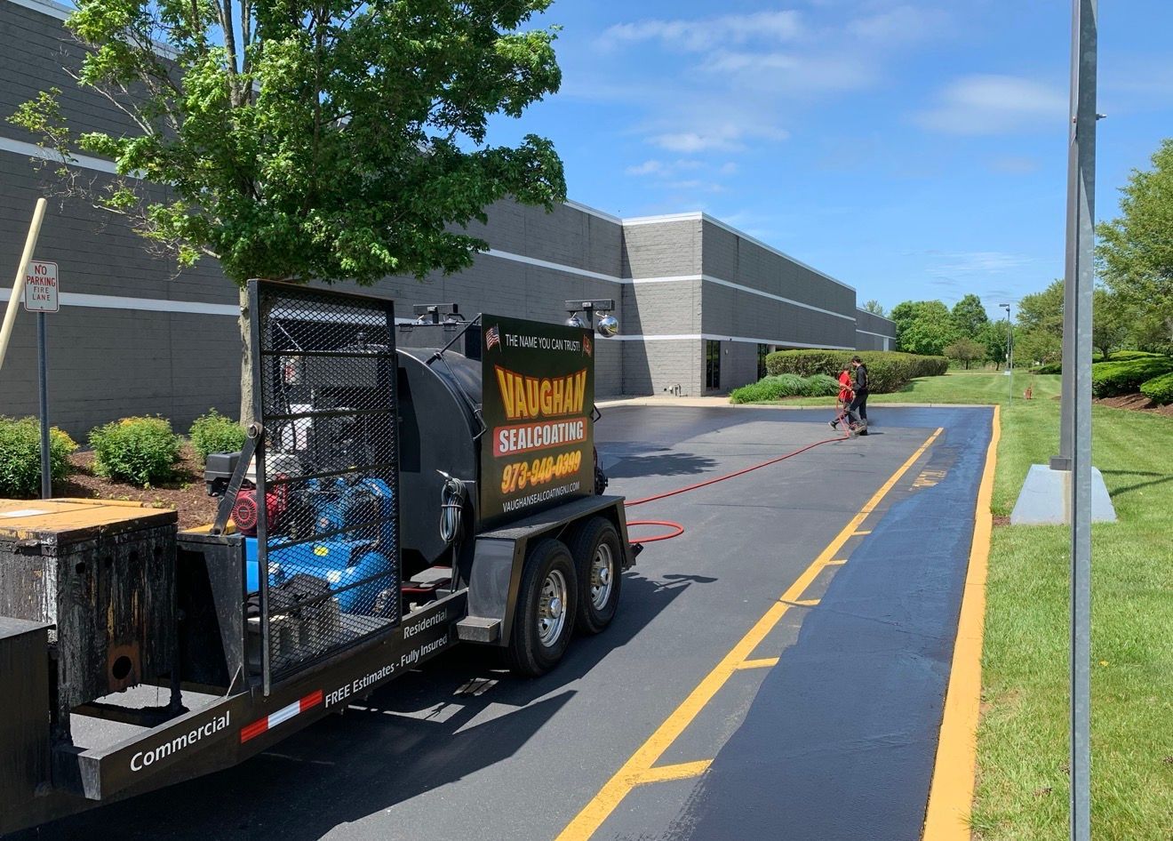 A worker operates a trailer sprayer to apply black sealant to an asphalt parking lot next to a large grey building.
