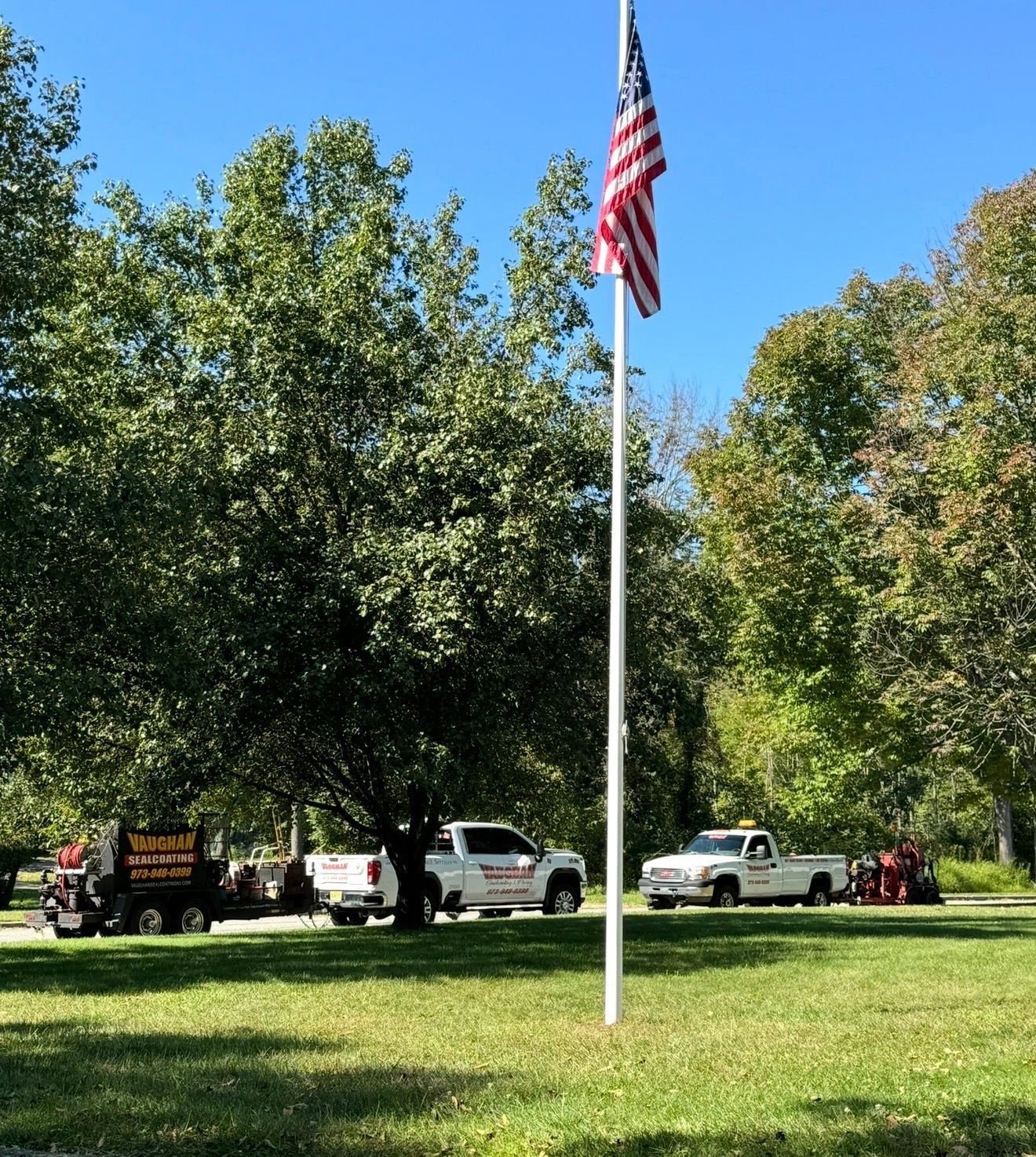 An American flag flies on a pole in a grassy park, with two white work trucks parked in the background under large trees.