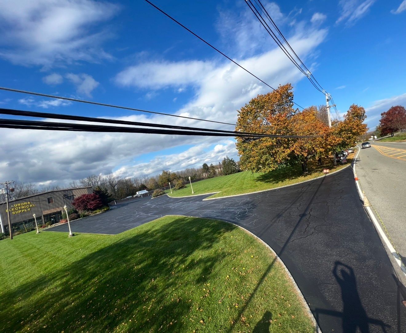 Newly paved asphalt driveway extending from a suburban road into a grassy yard with trees under a blue sky.