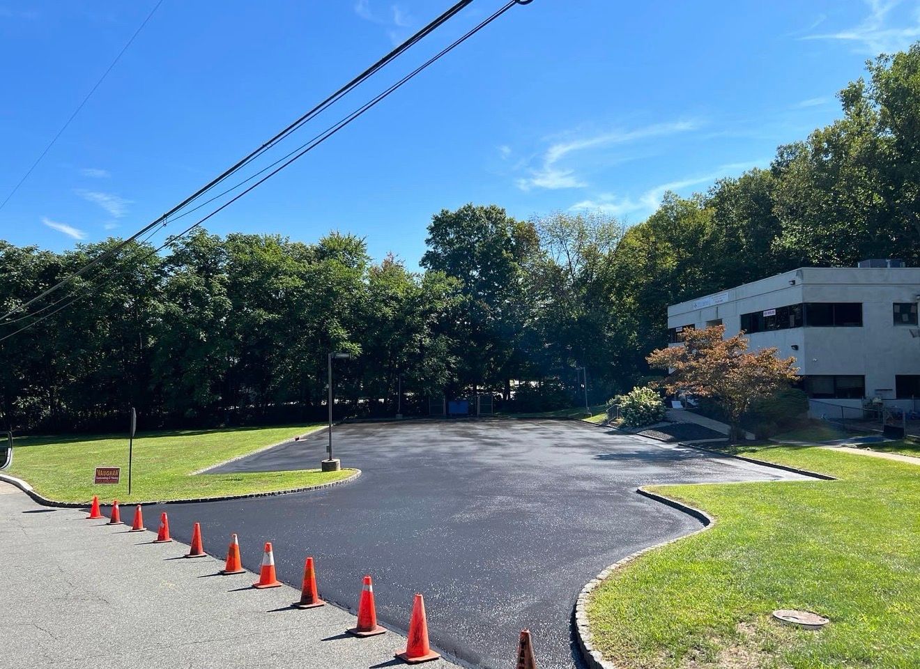 A freshly paved asphalt parking area lined with orange safety cones, situated next to a light-colored building and trees.