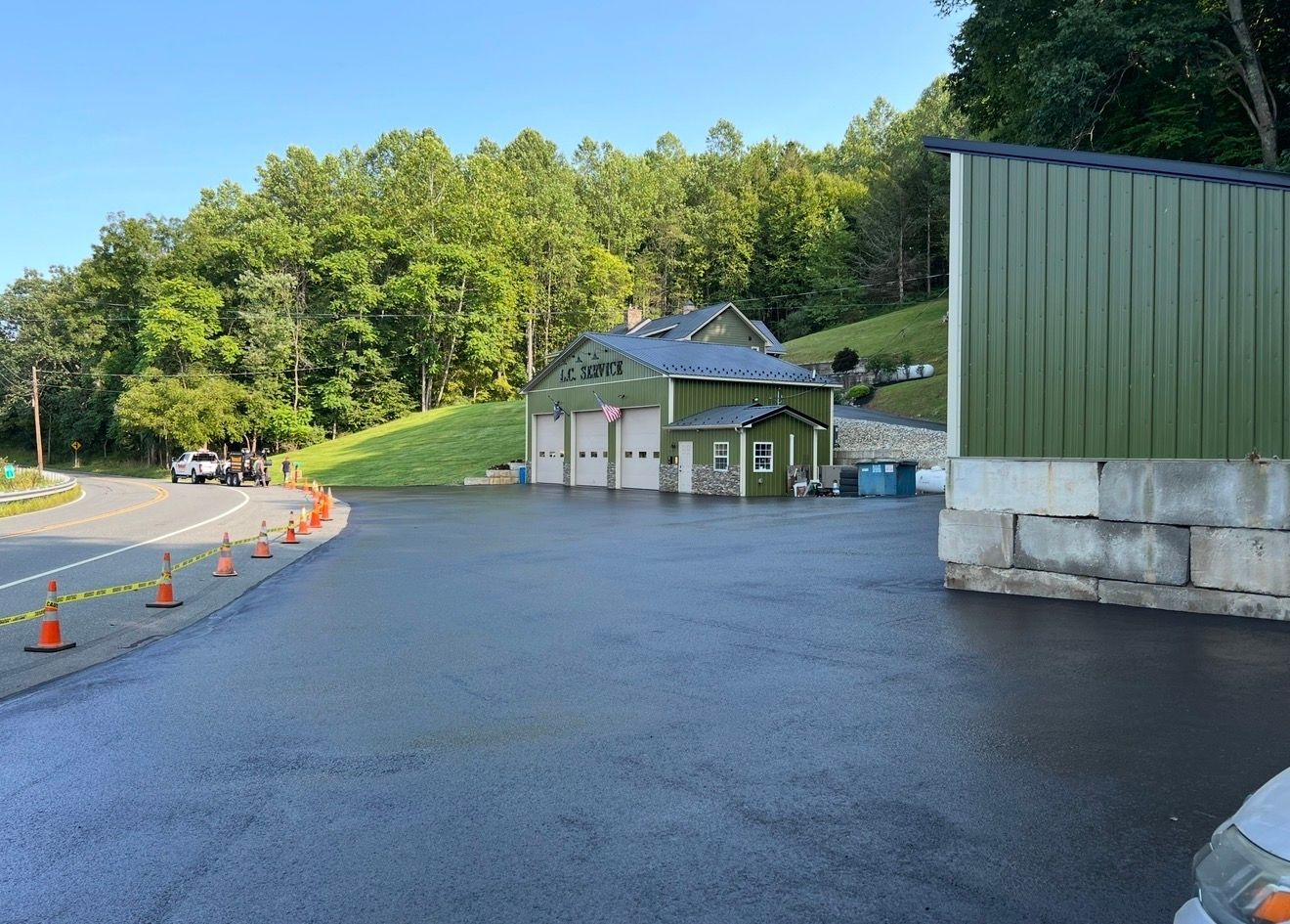 A green fire station with three garage bays sits at the base of a forested hill, beside a paved lot with safety cones.