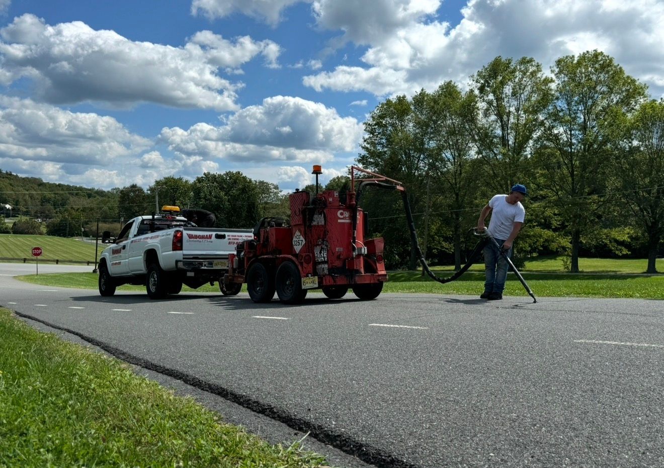 A worker uses a hose from a red tow-behind machine connected to a white pickup truck on an asphalt road.