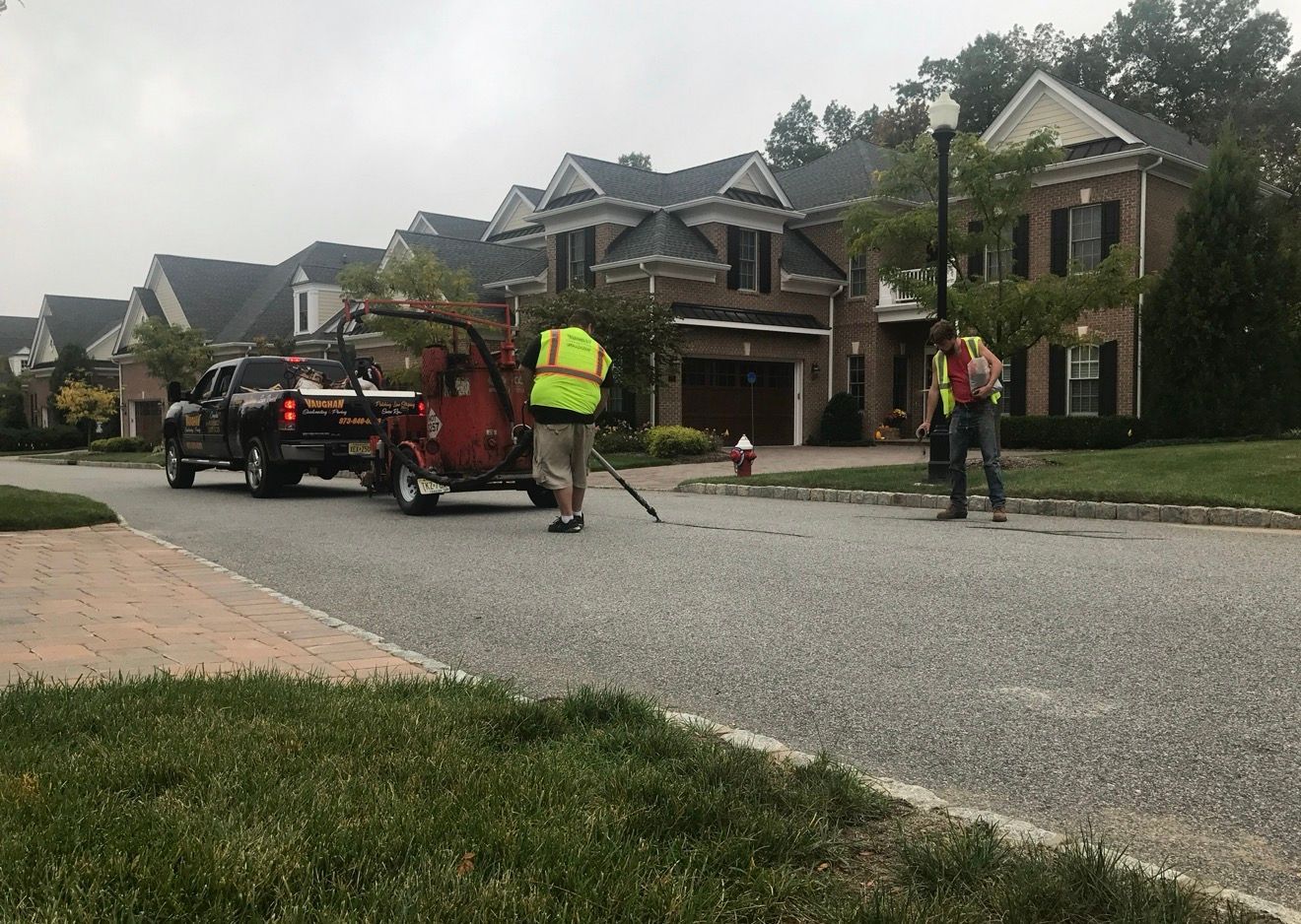 Two workers in high-visibility vests operate equipment on a residential street in front of large brick homes.