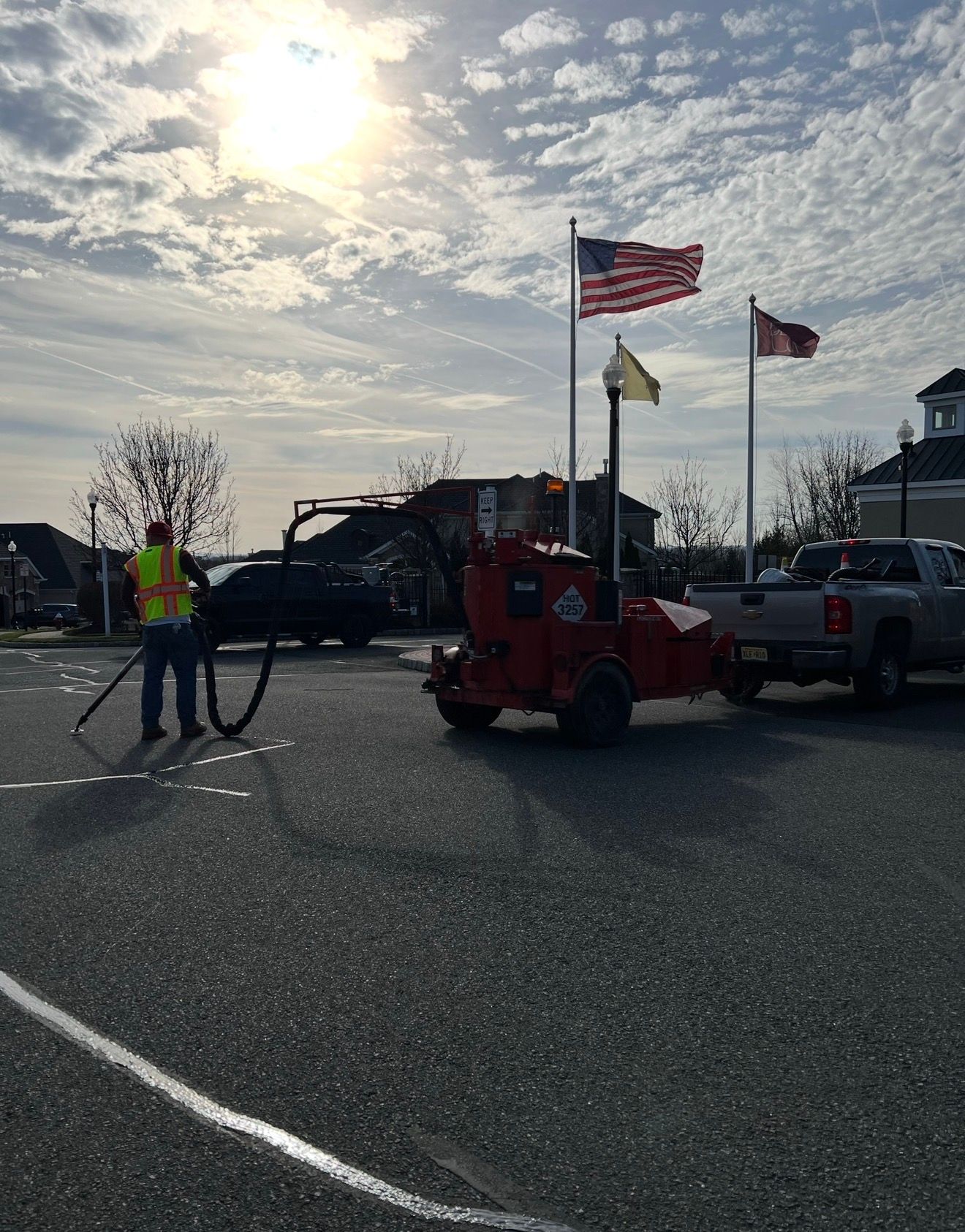 A worker in a high-visibility vest operates equipment to seal cracks in a parking lot under a bright sun and flags.