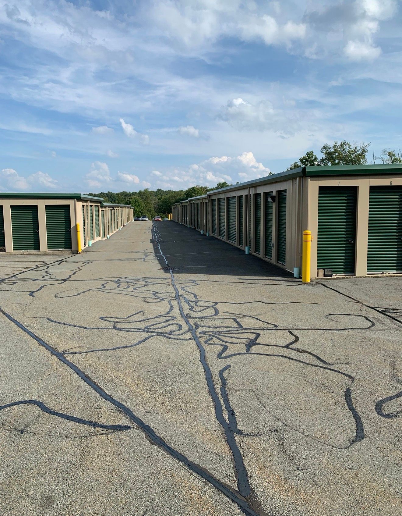 Rows of tan self-storage units with green roll-up doors line a gravel path under a partly cloudy sky.