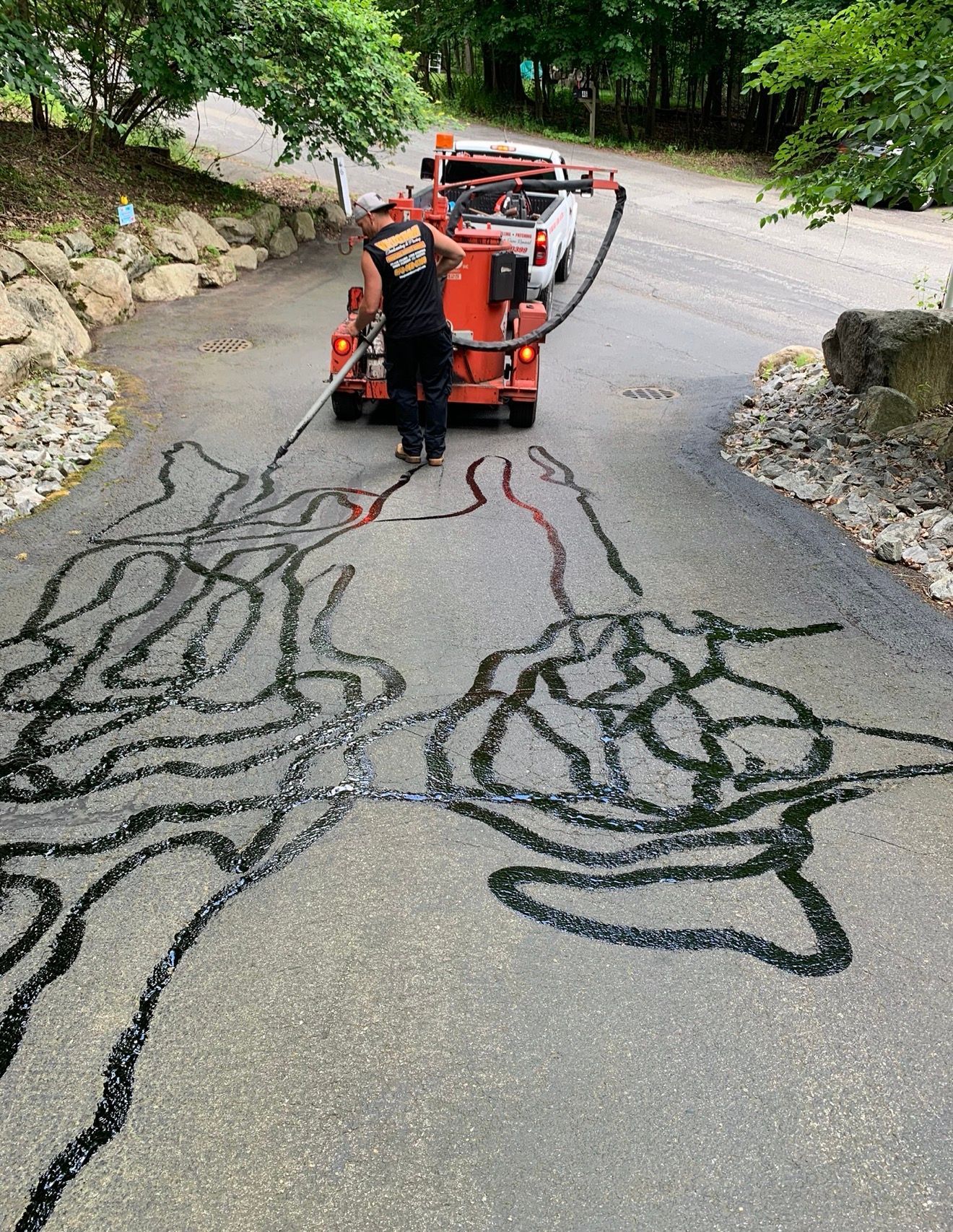 A worker uses a crack-sealing machine to fill large, hand-shaped designs on a paved driveway surrounded by trees.