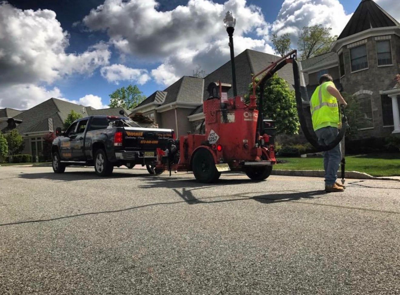 A worker in a high-visibility vest uses a red asphalt crack sealing machine towed by a pickup truck on a residential street.