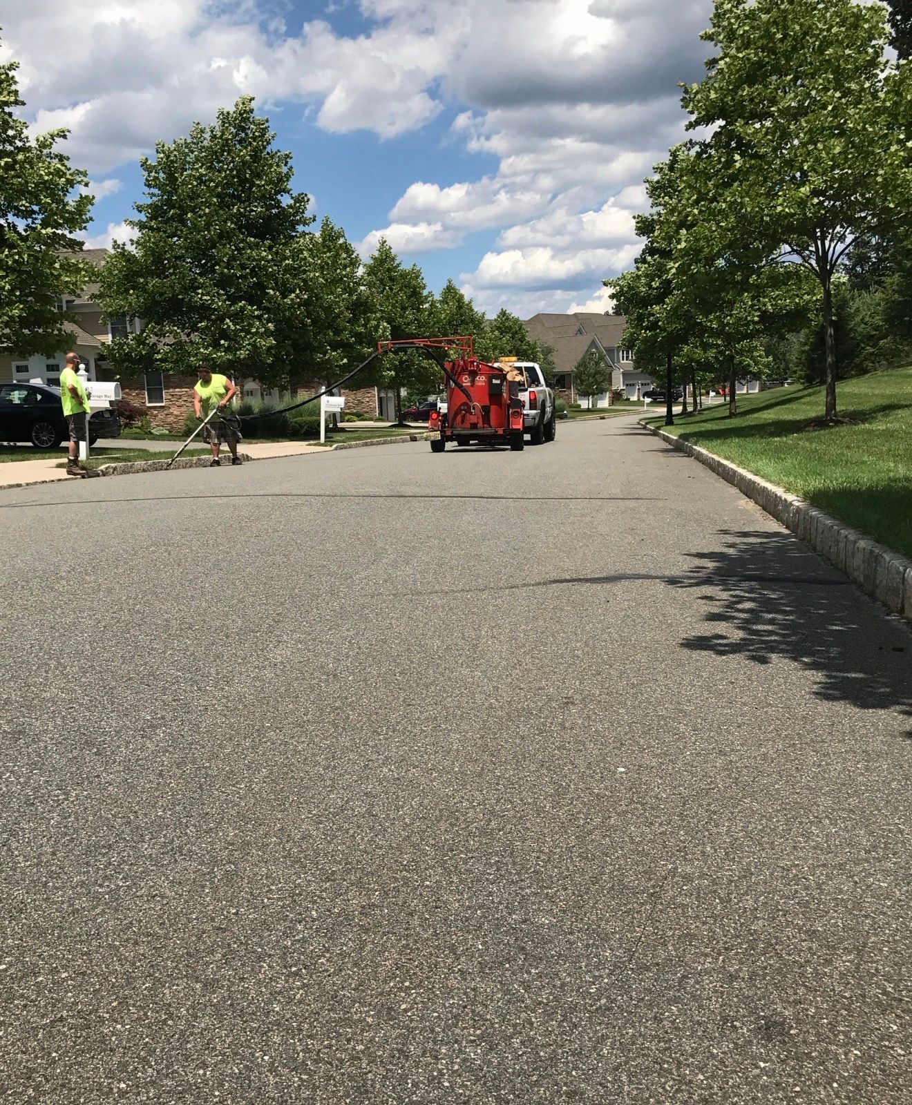 Workers in high-visibility vests operate a wood chipper on a suburban street lined with trees under a cloudy sky.