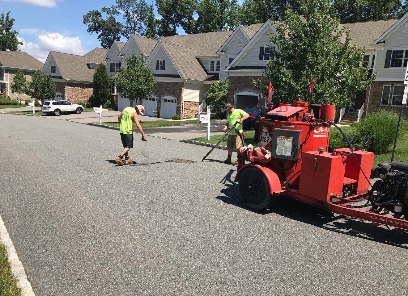 Two workers in neon safety vests perform asphalt crack sealing on a residential street using a red trailer-mounted melter.