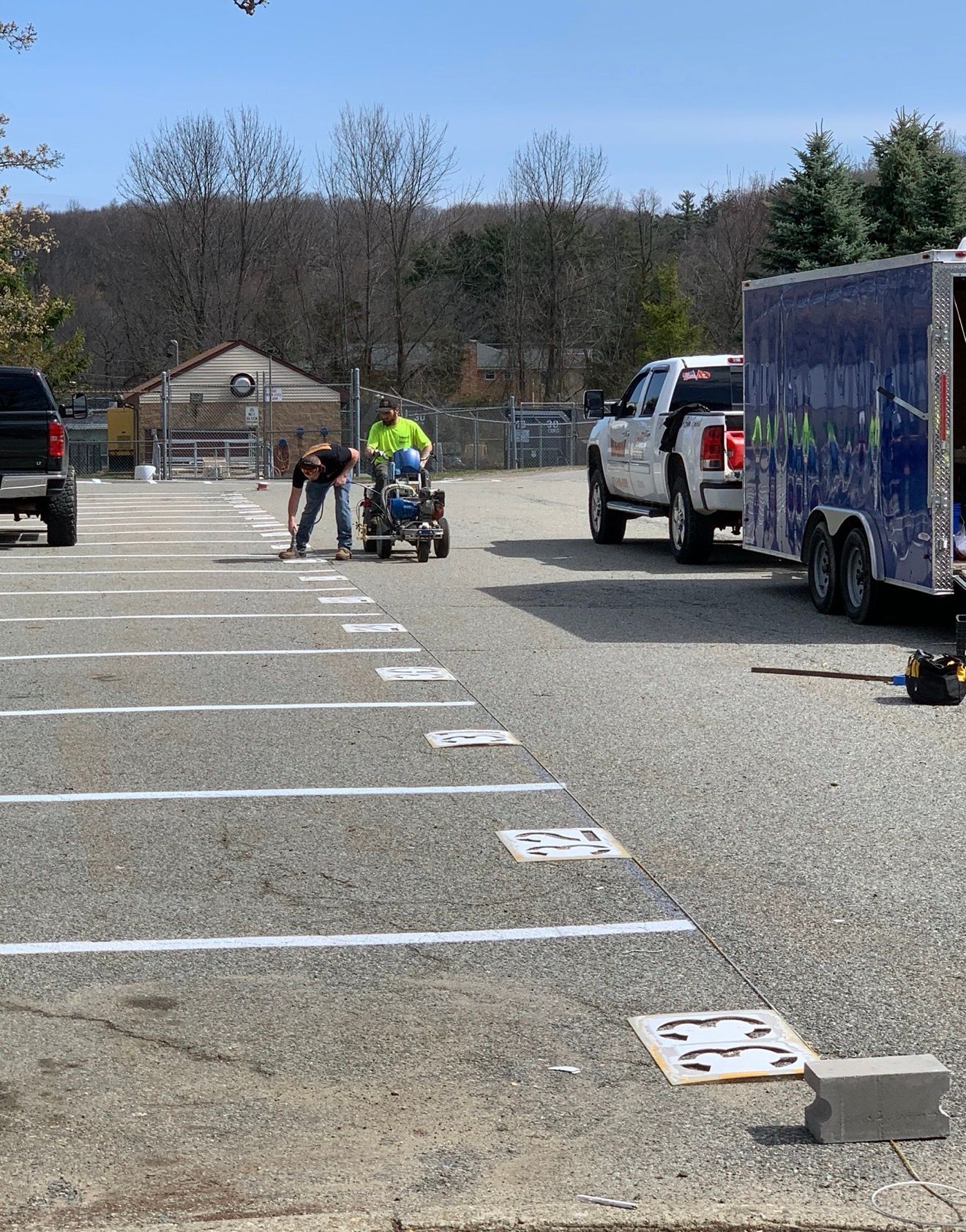 Two people use a machine to paint white parking space lines and numbers on a gravel lot next to a truck and trailer.