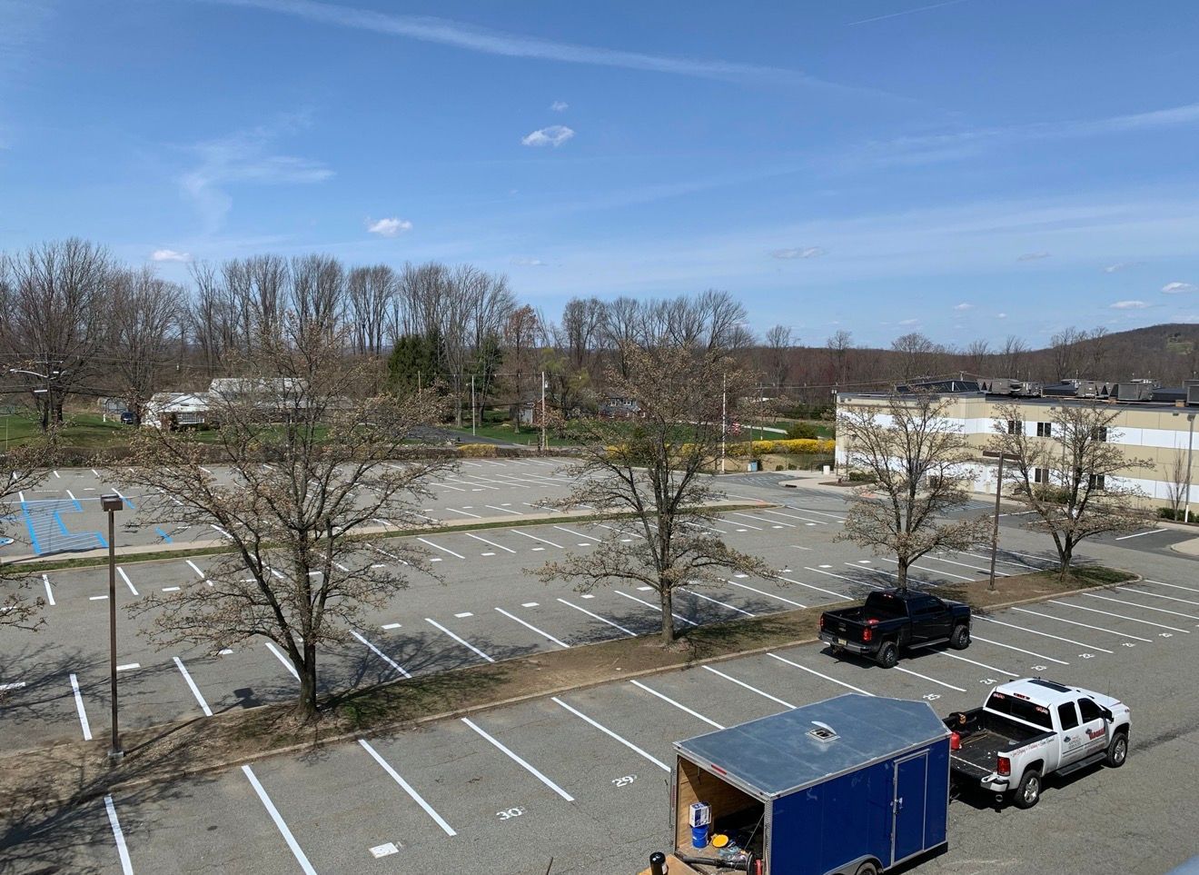 A wide, outdoor parking lot under a blue sky, featuring bare trees, a parked black truck, and a trailer with a white truck.