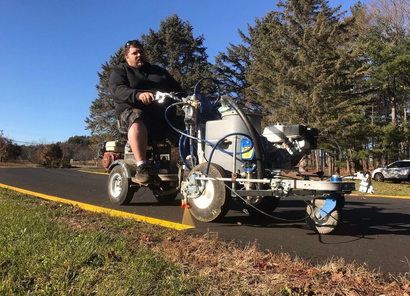 A person rides a motorized line-striping machine, painting a yellow line along the edge of a paved road.
