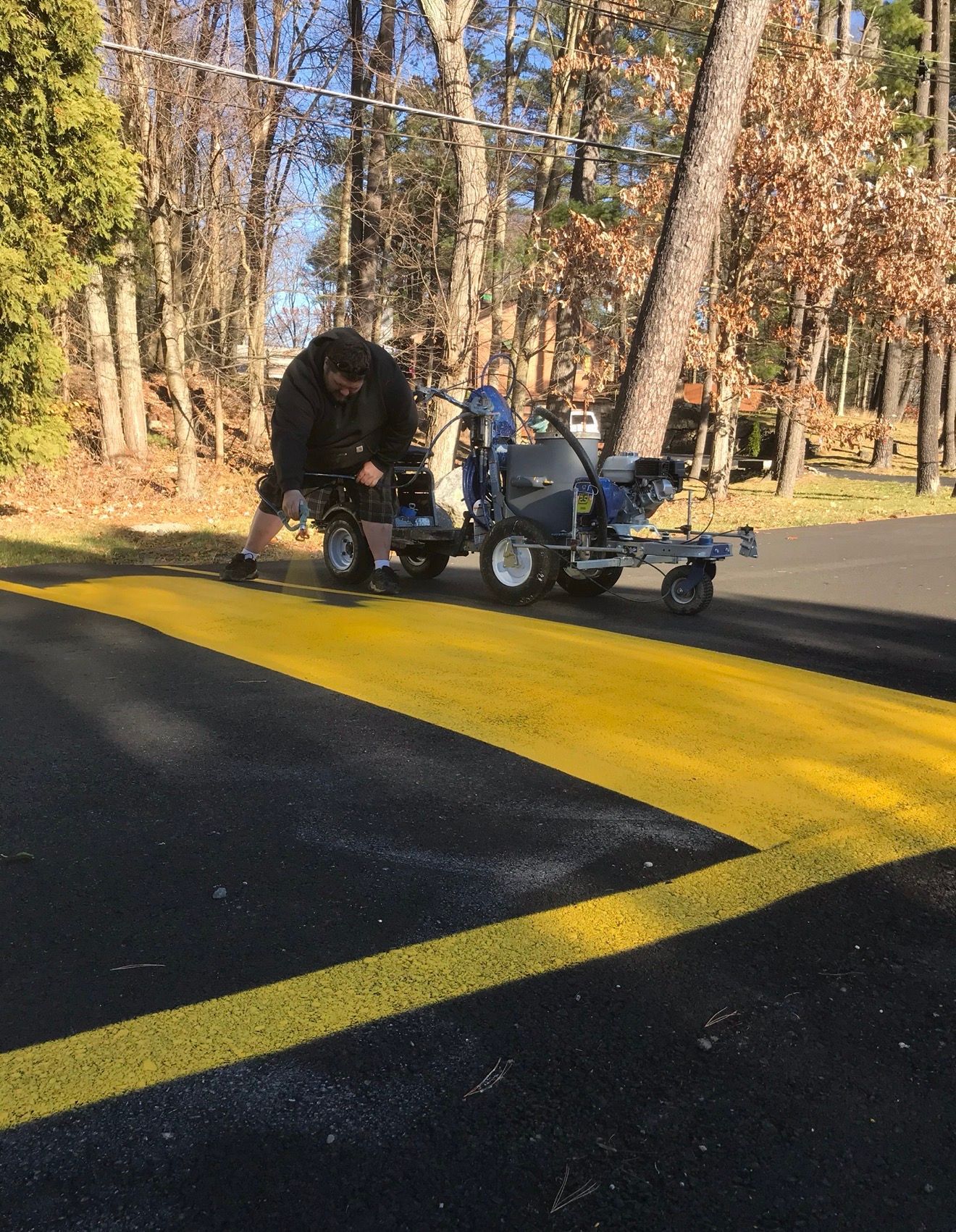 A person uses a walk-behind line striping machine to paint a wide yellow stripe on fresh black asphalt outdoors.